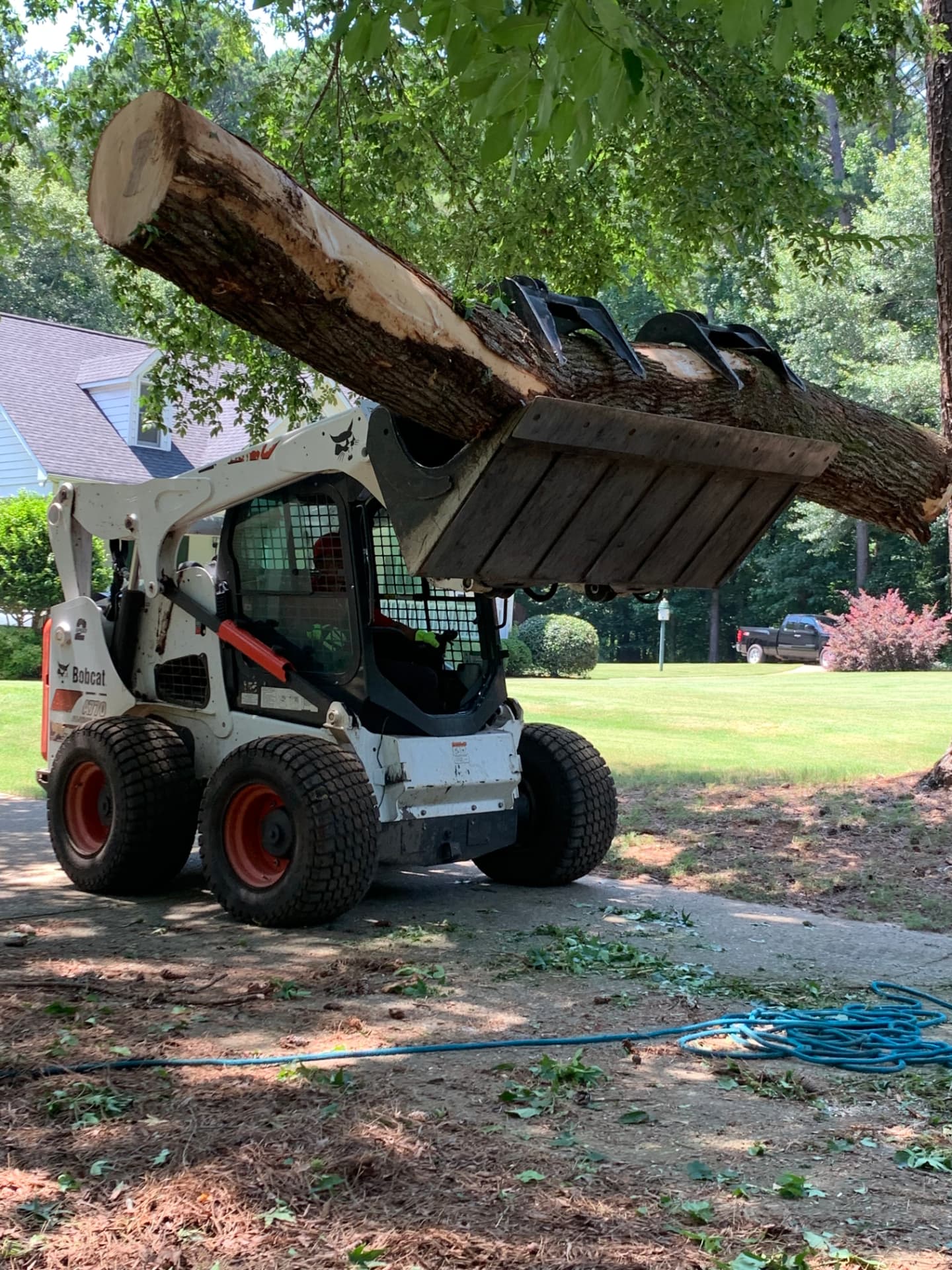 Skid-steer loader with grapple lifting a large tree log on a residential driveway